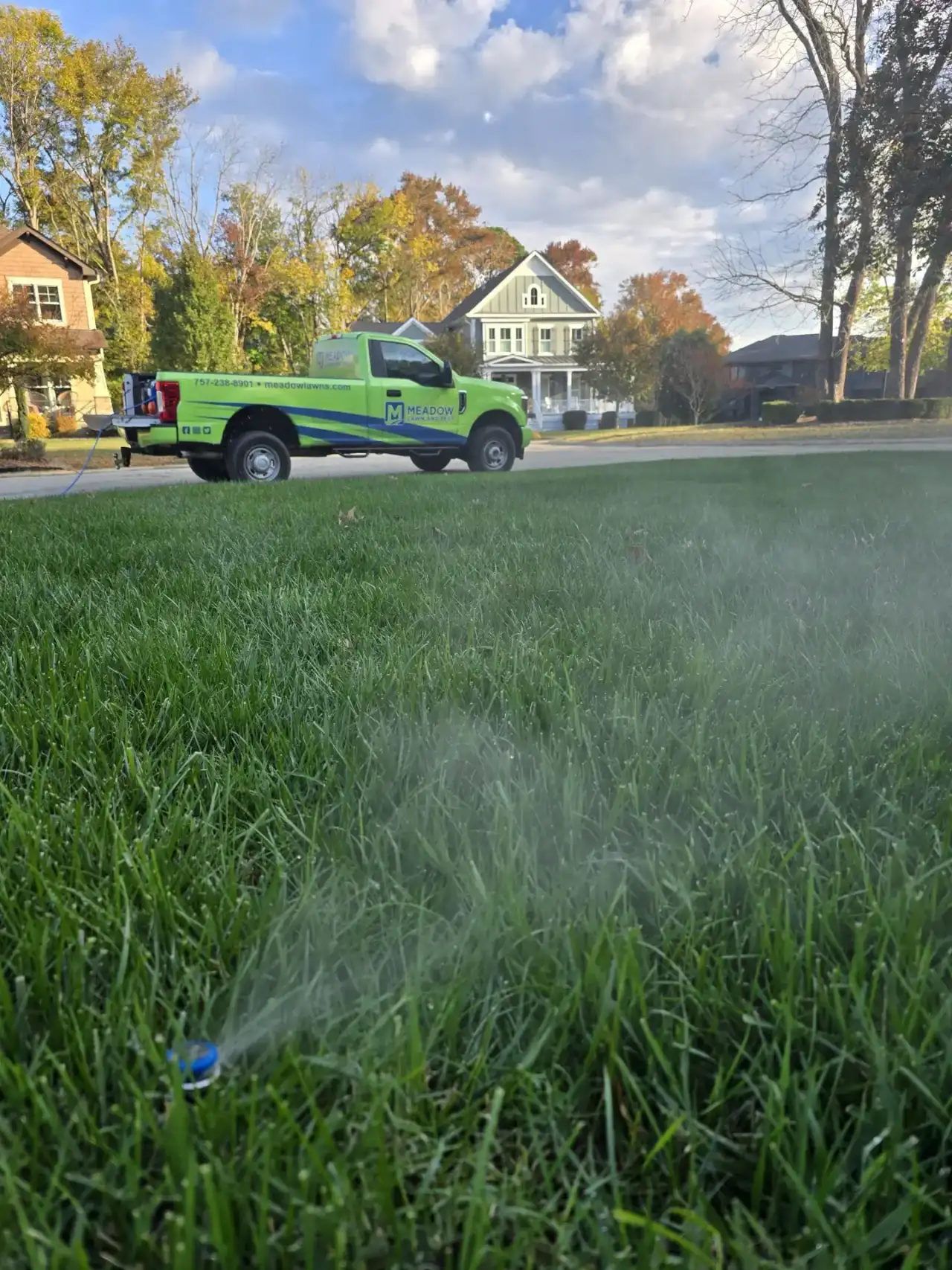Sprinkler head watering green lawn with Meadow Lawn & Pest truck parked curbside in Carrollton neighborhood