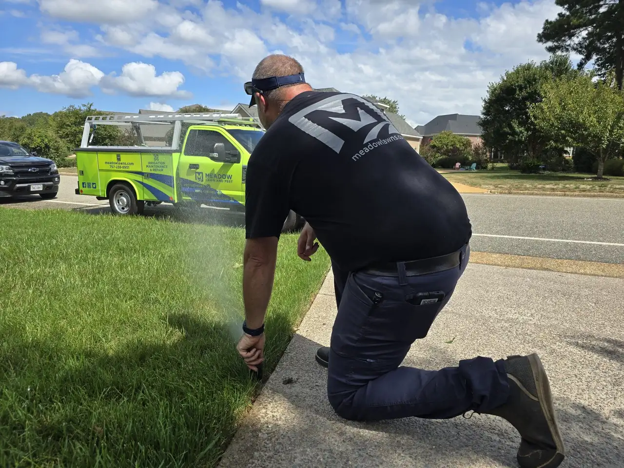 Technician kneeling to adjust a spraying sprinkler head beside service truck in Carrollton