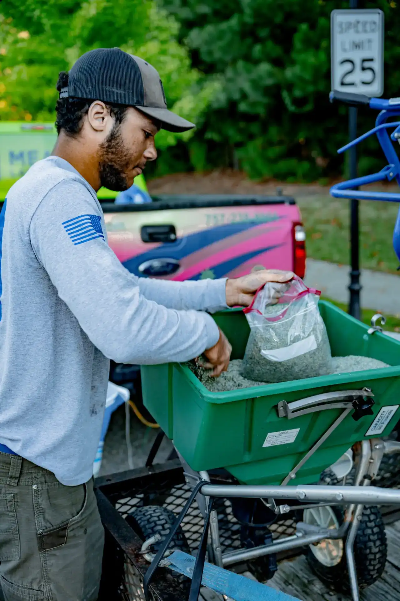 Lawn care technician filling a green broadcast spreader with granular fertilizer beside a work truck